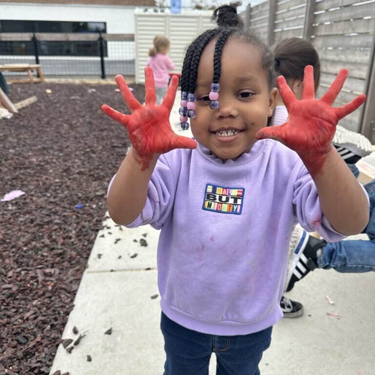 A toddler smiles and holds up her hands, which are covered in red paint.